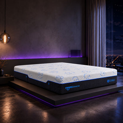 מזרן Roomi Dreamer SmartBlocks® עם חלק עליון לבן מעוטר ודפנות כהות, מונח על מיטה מודרנית בשעת לילה.